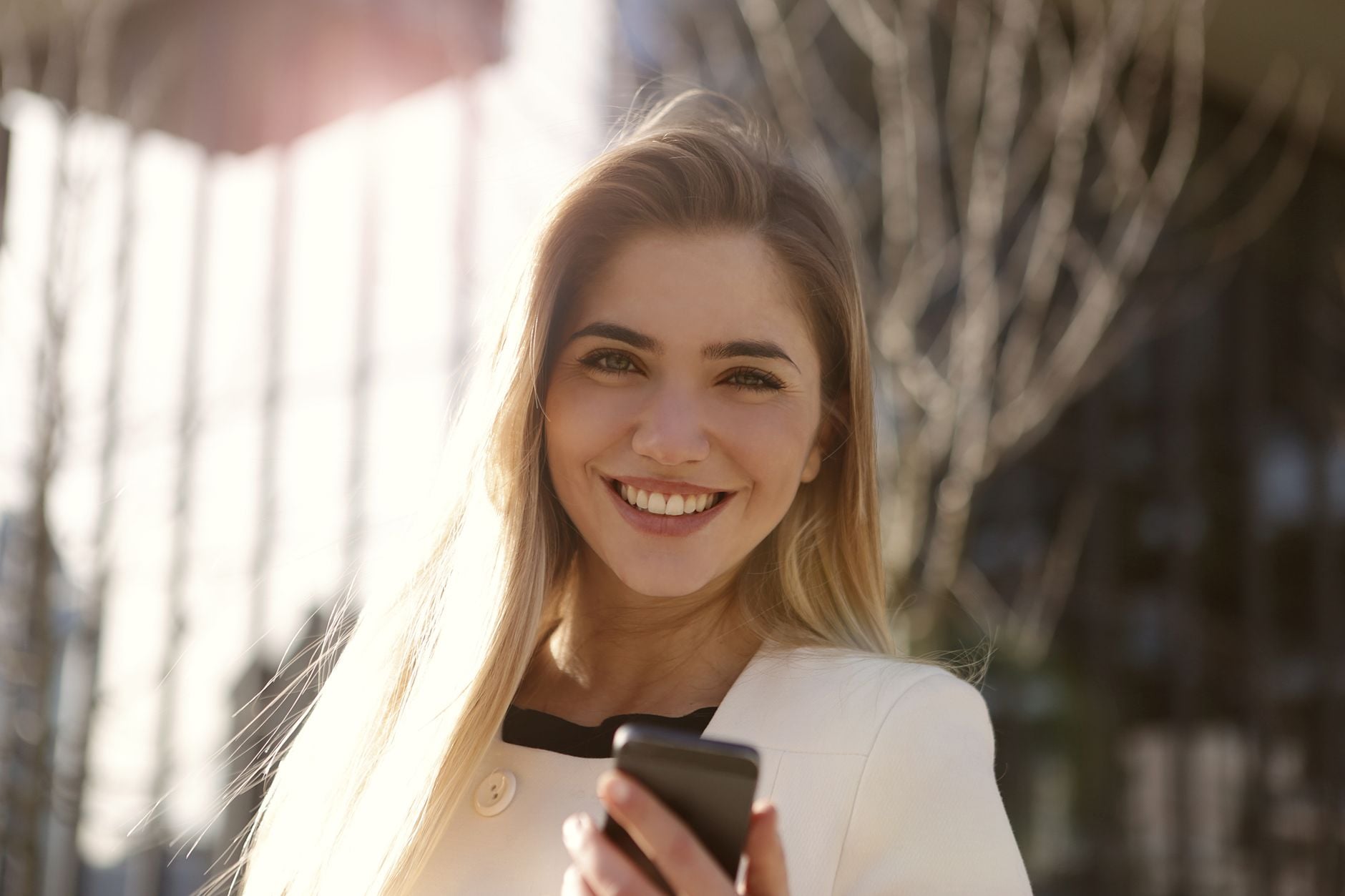 Donna sorridente con capelli castani che tiene uno smartphone in una strada urbana con edifici sullo sfondo