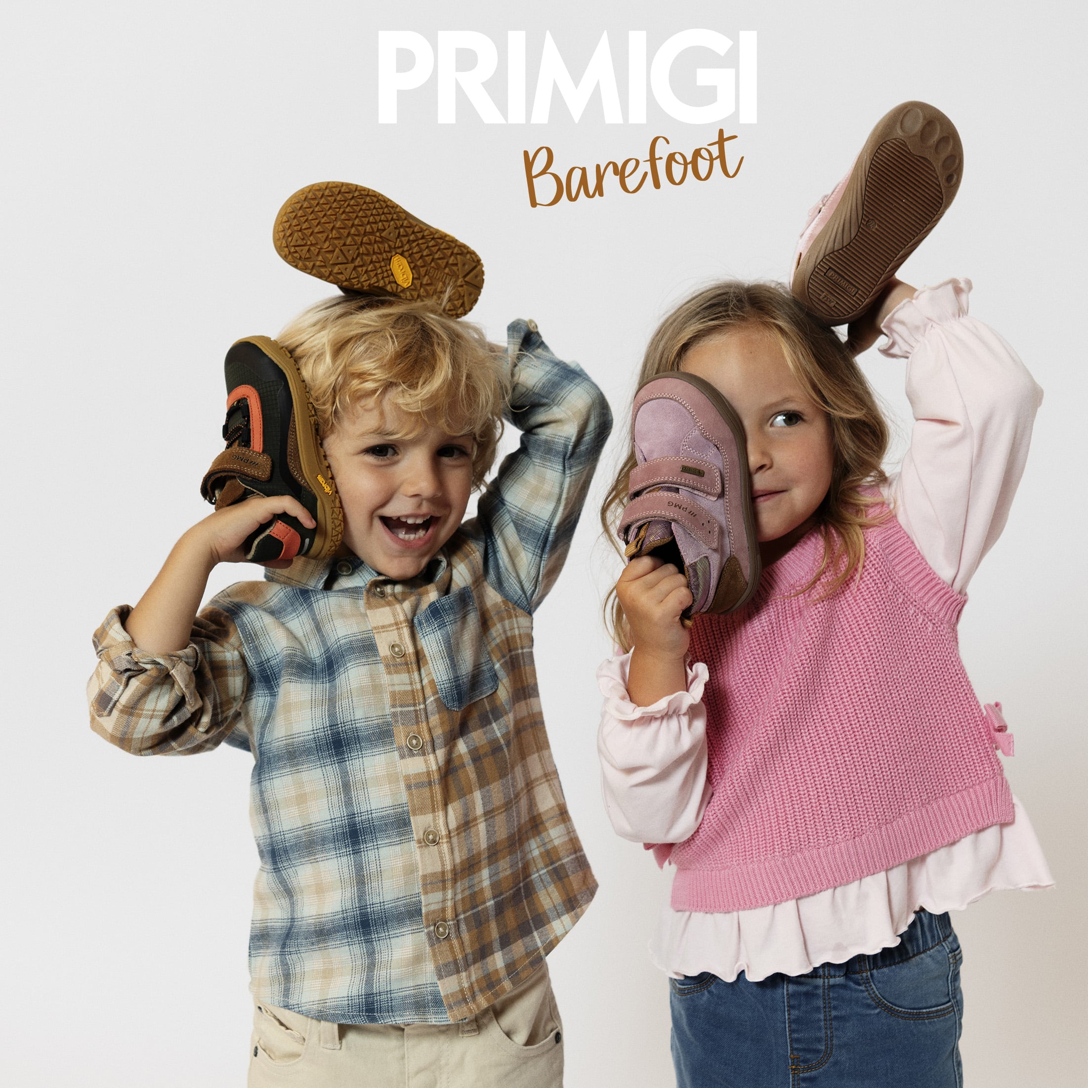 Due bambini sorridenti con scarpe barefoot Primigi, uno con camicia scozzese e uno con maglione rosa.