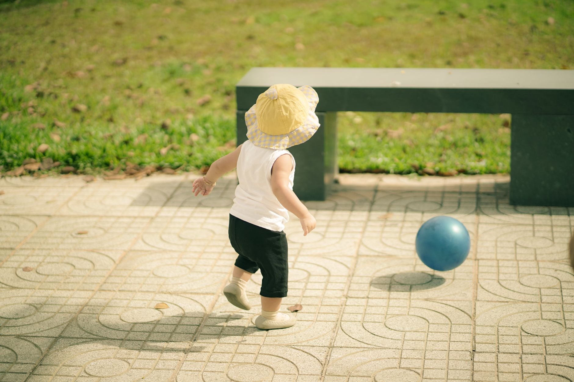 Bambino piccolo in cappello giallo corre verso una palla blu su una pavimentazione geometrica in un giardino