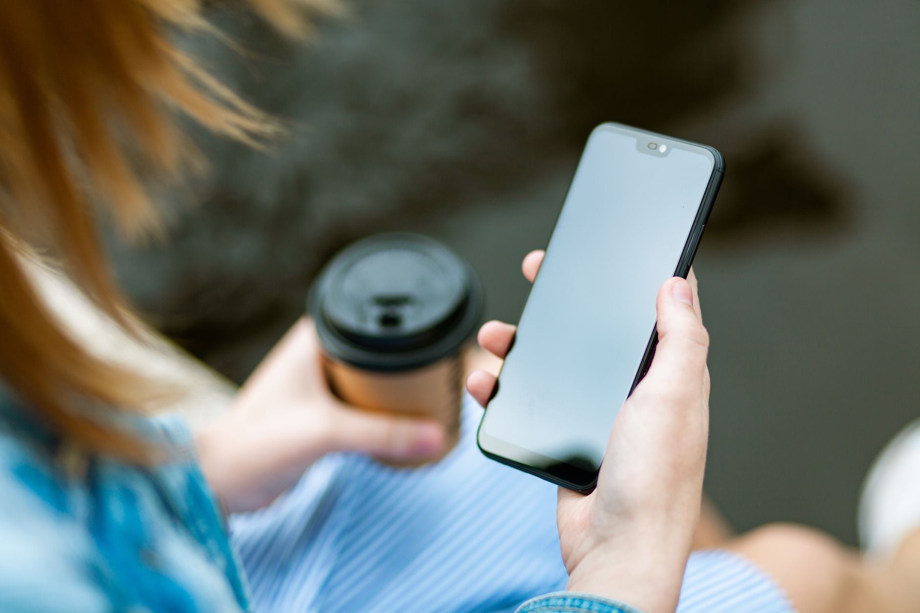 Donna con capelli biondi che tiene uno smartphone con schermo bianco e un caffè in mano