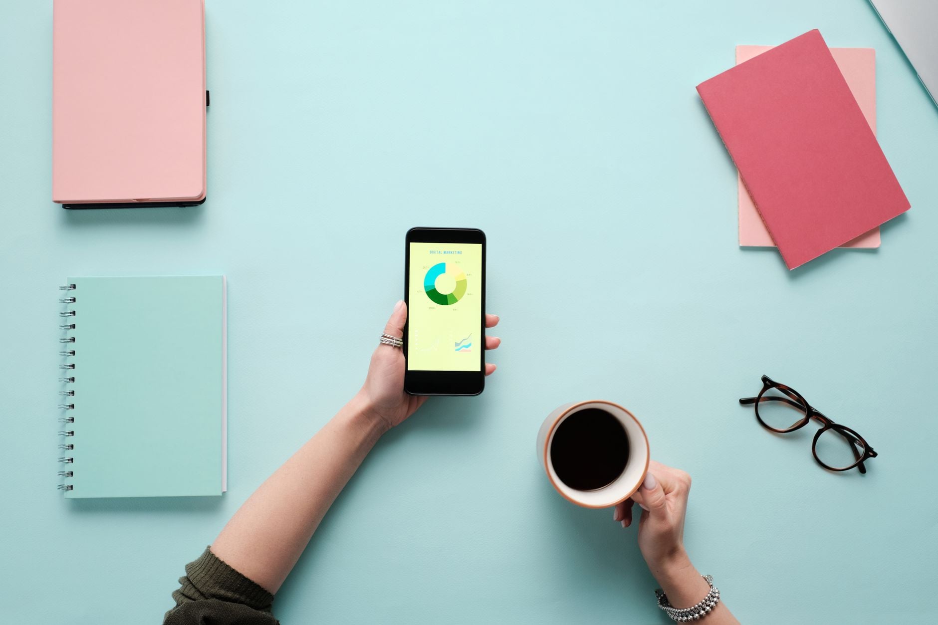 Mano regge smartphone con grafico colorato circolare e tazza di caffè su scrivania azzurra con quaderni e occhiali