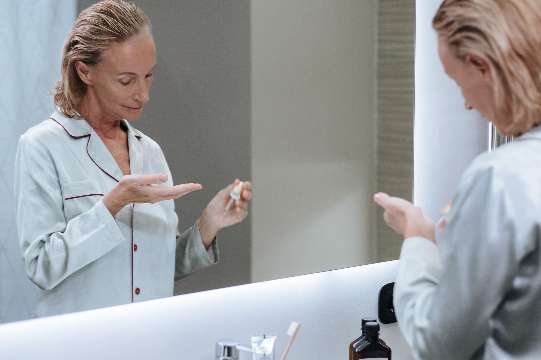 Donna in camice bianco osserva il riflesso allo specchio mentre tiene un prodotto di skincare in mano