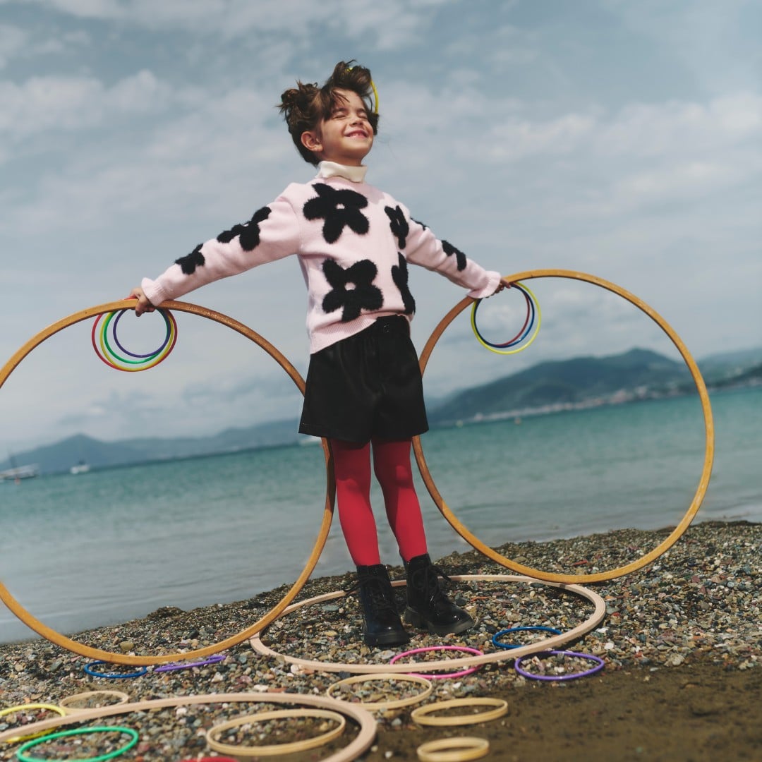 Bambino in maglione rosa con disegni neri e calzamaglia rossa che gioca con hula hoop colorati sulla spiaggia.