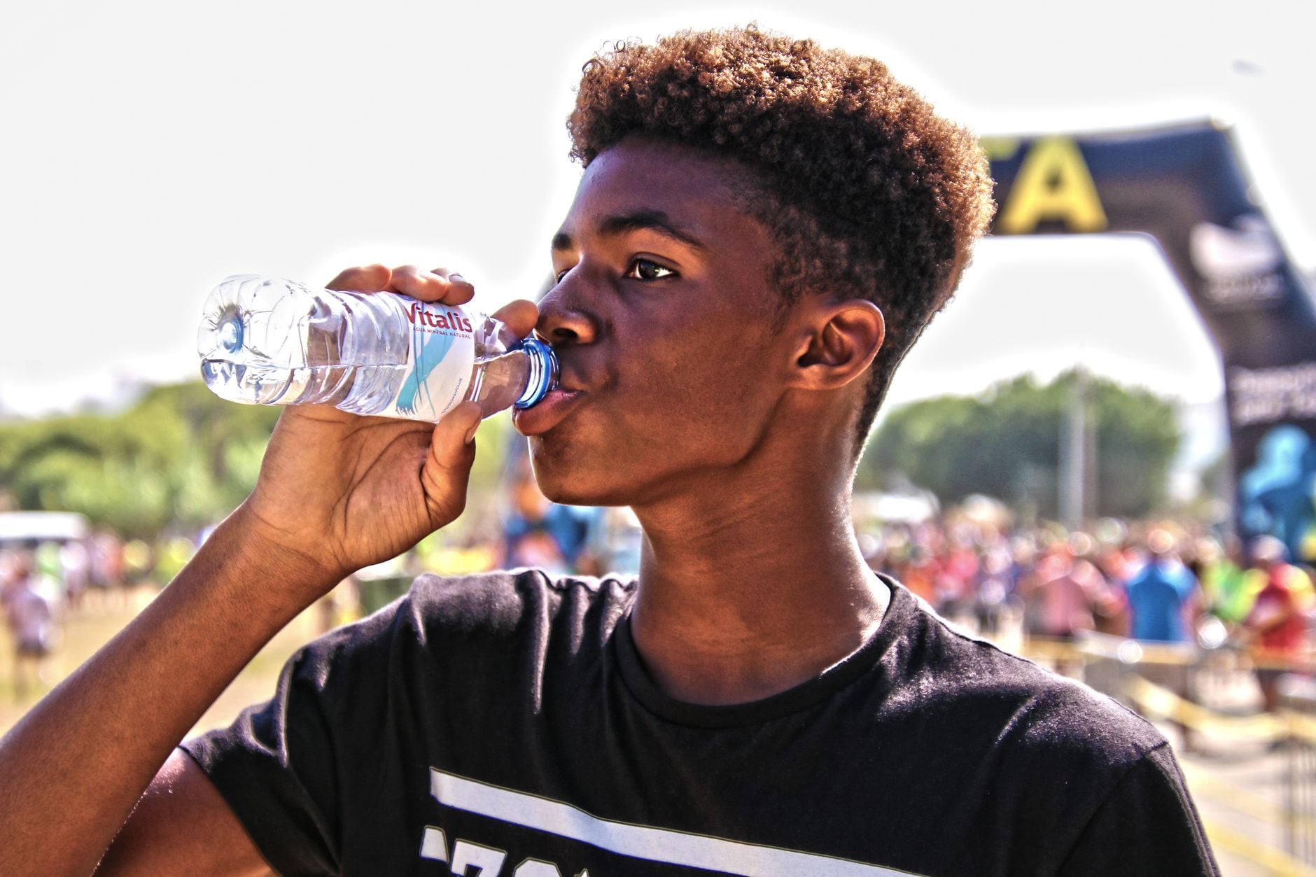Giovane atleta beve da una bottiglia d'acqua durante una gara all'aperto con folla sullo sfondo