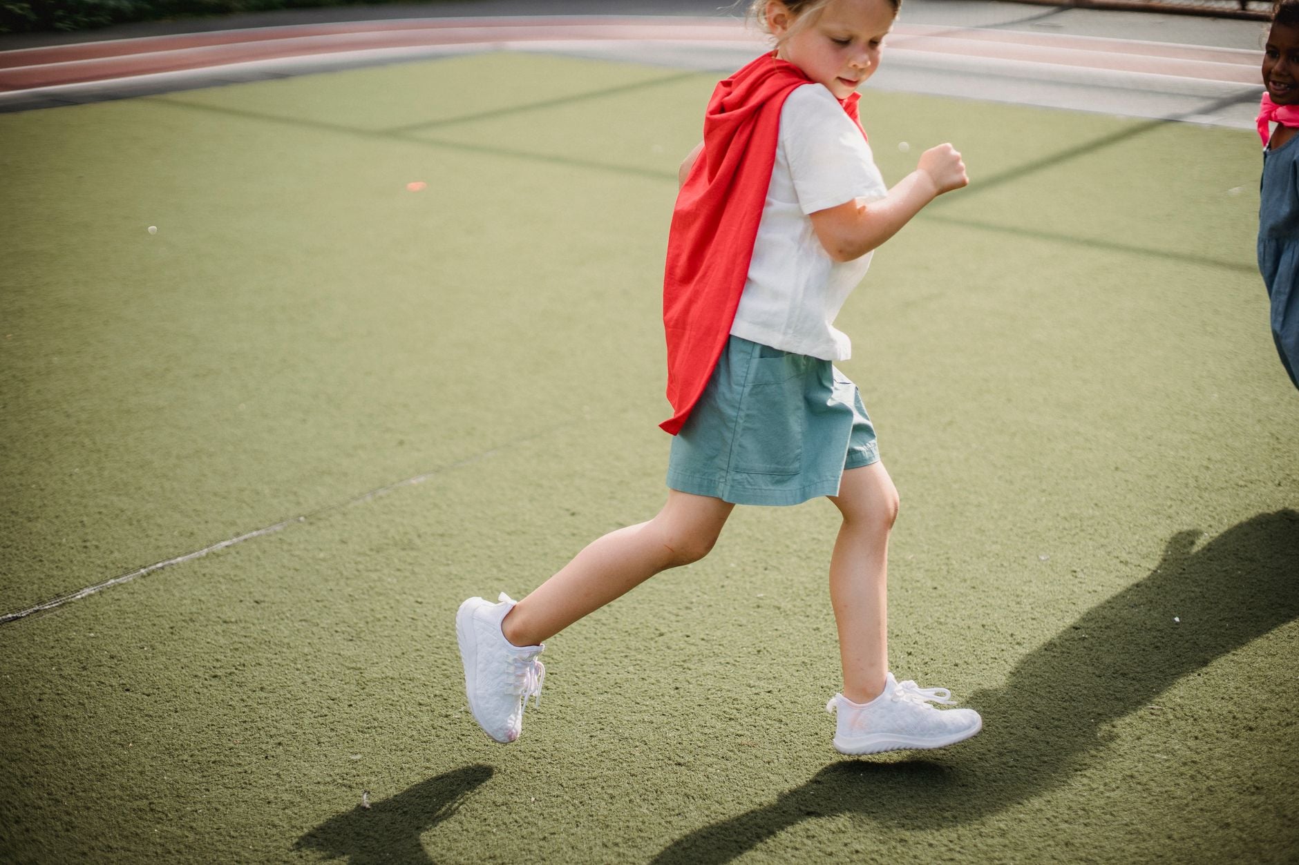 Bambina che corre su un campo da tennis indoor indossando un mantello rosso e scarpe da ginnastica bianche