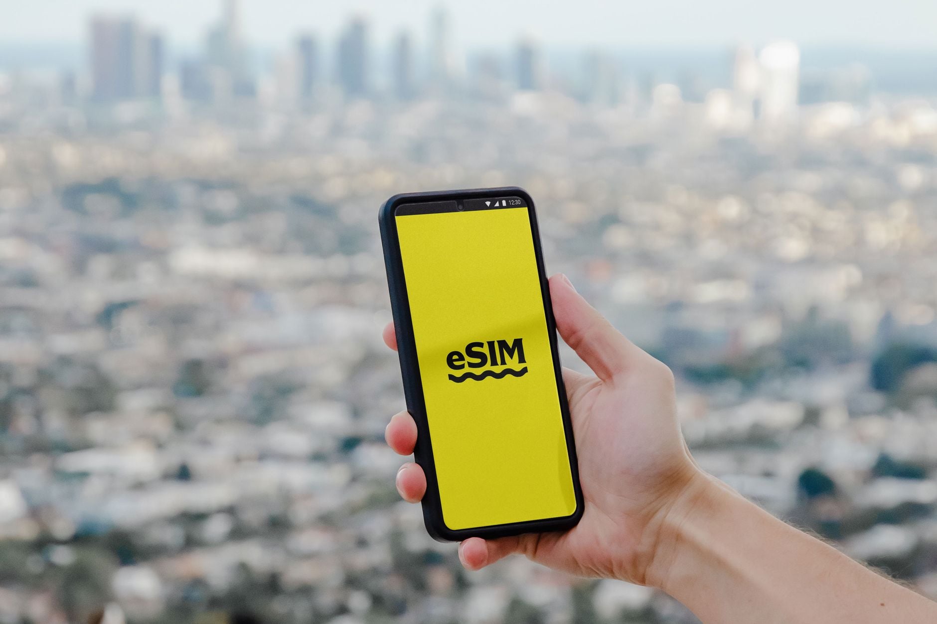 Mano che tiene uno smartphone nero con schermo giallo che mostra logo eSIM, sfondo città sfocata